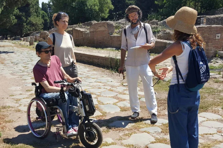 Taşlı tarihi mekanda tekerlekli sandalye kullanıcısı erkek turist ve yanında 2 kadın 1 erkek olan ayakta sohbet ediyorlar.