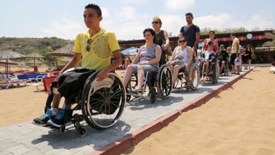 Bu fotoğrafta, tekerlekli sandalye kullanan bireyler sahil kenarında özel olarak döşenmiş bir yolda ilerliyor. Kumsalın ortasından geçen bu taş döşeli yol, engelli bireylerin plaja rahatça ulaşabilmesi için yapılmış. Ön sırada genç bir erkek gülümsüyor; arkasında kadınlar ve erkeklerden oluşan bir grup var. Bazı bireyler kendi sandalyelerini sürerken, bazılarına refakat eden kişiler eşlik ediyor. Arka planda şemsiye ve şezlonglar, ayrıca sahil kafesi benzeri bir yapı görülüyor.