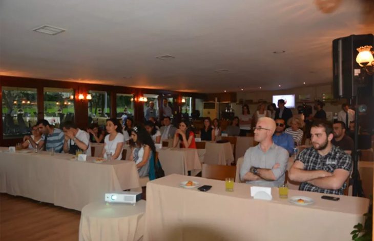 Bu fotoğrafta, kapalı bir salonda düzenlenen bir toplantı ya da seminer etkinliği görülüyor. Katılımcılar masalara yerleştirilmiş ve çoğu dikkatle öne bakıyor; önde bir projeksiyon cihazı da bulunuyor, bu da bir sunum yapıldığını veya yapılmakta olduğunu gösteriyor. Masalarda meyve suyu, atıştırmalıklar ve peçetelikler yer almakta. Arka planda ise ayakta bekleyen ya da sohbet eden katılımcılar var.