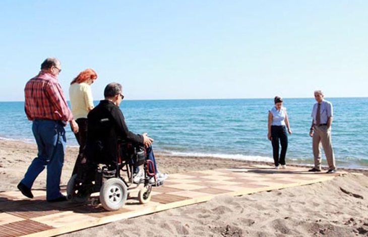 Bu fotoğrafta, kumsalda yerleştirilmiş ahşap bir yürüyüş yolunda yürüyen ve tekerlekli sandalye kullanan bireyler görülüyor. Yol, kumun üstüne serilerek hem engelli bireylerin hem de yaşlı ya da hareket kabiliyeti sınırlı kişilerin denize ulaşımını kolaylaştıracak şekilde tasarlanmış. Ön planda bir kişi elektrikli tekerlekli sandalyesiyle ilerliyor; yanında ona eşlik eden kişiler var. Arka planda ise deniz kenarında duran iki kişi görülüyor. Hava açık ve güneşli, deniz ise sakin.