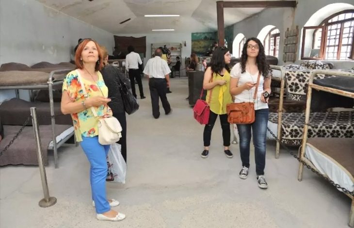 Bu fotoğrafta, bir grup insan bir müzeyi ya da tarihi bir yapıyı ziyaret ediyor. Mekân, eski tarz ranzalarla dolu geniş bir oda; bu da buranın geçmişte yatakhane olarak kullanıldığını düşündürüyor. Ranzalarda battaniyeler serili, çoğu metal iskeletli ve üst üste yerleştirilmiş. Ziyaretçiler dikkatle etrafı inceliyor, ellerinde rehber kitapçıklar veya telefonlar bulunuyor. Arka planda duvarda bir tablo ve bazı eski eşyalar görülüyor.