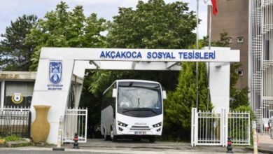 Bu fotoğrafta, Ankara Büyükşehir Belediyesi'ne ait Akçakoca Sosyal Tesislerinin giriş kapısı görülüyor. Kapının hemen altında bir servis otobüsü (Isuzu marka) park etmiş durumda. Girişin üst kısmında tesisin ismi mavi harflerle yazılmış ve sağ üst köşede Türk bayrağı dalgalanıyor. Girişin yanında, belediyeye ait bir güvenlik kulübesi ve tesisin logosu yer almakta. Arka planda yeşillikler ve tesis binası görünmekte.