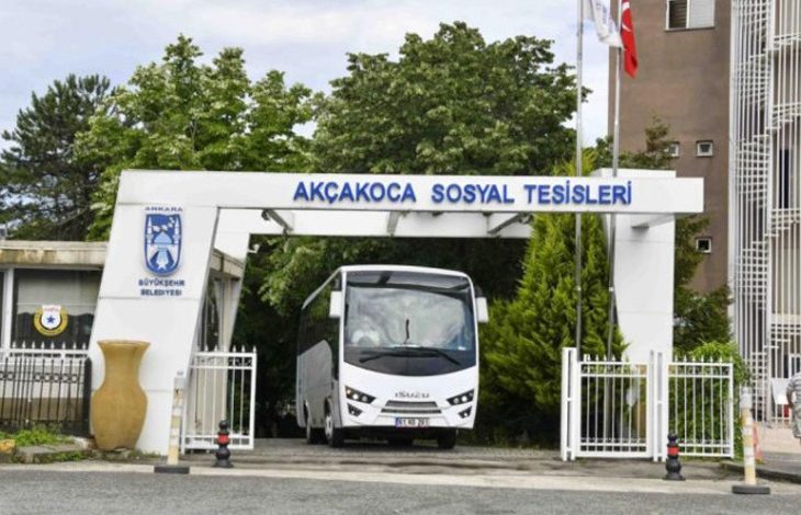 Bu fotoğrafta, Ankara Büyükşehir Belediyesi'ne ait Akçakoca Sosyal Tesislerinin giriş kapısı görülüyor. Kapının hemen altında bir servis otobüsü (Isuzu marka) park etmiş durumda. Girişin üst kısmında tesisin ismi mavi harflerle yazılmış ve sağ üst köşede Türk bayrağı dalgalanıyor. Girişin yanında, belediyeye ait bir güvenlik kulübesi ve tesisin logosu yer almakta. Arka planda yeşillikler ve tesis binası görünmekte.