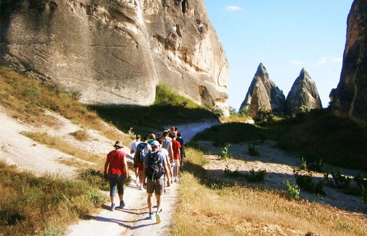 ChatGPT: Bu fotoğrafta, doğa yürüyüşü (trekking) yapan bir grup insan, Kapadokya bölgesine özgü peribacalarının arasında yürüyüş yaparken görülüyor. Katılımcılar sırt çantaları ve şapkalarıyla donanmış, patika bir yolda sırayla ilerliyorlar. Etrafta yüksek kaya oluşumları ve sarı-yeşil tonlarda doğal bitki örtüsü göze çarpıyor