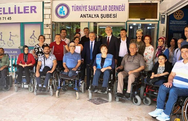 Bu fotoğrafta, Türkiye Sakatlar Derneği Antalya Şubesi önünde toplanmış bir grup insan görülüyor. Ön sırada çoğunluğu tekerlekli sandalye kullanan bireyler yer almakta; arkada ise ayakta duran destekçiler, dernek yetkilileri ve gönüllüler bulunuyor. Çeşitli yaş gruplarından kadın ve erkekler bir araya gelmiş, birlik ve dayanışma duygusu güçlü bir şekilde yansıtılmış. Arka planda derneğin tabelası ve “Aktif Engelliler” yazısı yer alıyor.