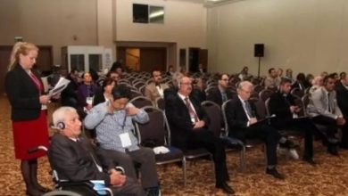 Bu fotoğraf, büyük bir konferans veya seminer salonunda gerçekleşen bir toplantıyı göstermektedir. Katılımcılar sıralı sandalyelerde oturmuş, bazıları not alıyor, bazıları dinliyor. Önde bir kadın ayakta konuşma yapıyor ya da sunum yapmaya hazırlanıyor. Önde tekerlekli sandalyede bir katılımcı dikkat çekiyor; bu da etkinliğin erişilebilir olduğunu ve engelli bireylerin de dahil edildiğini gösteriyor.