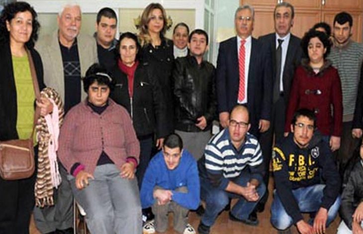 Bu fotoğraf, özel gereksinimli bireylerle birlikte çekilmiş bir toplu anı fotoğrafını göstermektedir. Fotoğrafta hem gençler hem de yetişkinler bulunuyor. Bazı bireylerin otizmli ya da zihinsel engelli bireyler olduğu gözlemlenebiliyor. Arkada takım elbiseli yetkililer ve öğretmen/veli gibi görünen bireyler yer almakta. Fotoğraf, bir ziyaret, etkinlik ya da farkındalık günü kapsamında çekilmiş olabilir. Ortam samimi ve destekleyici bir izlenim veriyor.