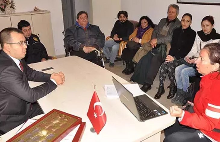 Bu fotoğraf, bir topluluk toplantısını veya kamuya açık bir görüşmeyi yansıtıyor. Masanın bir tarafında resmi kıyafetli bir yetkili otururken, diğer tarafında engelli bireyler ve destekçileri bulunuyor. Katılımcıların çoğu ciddi bir şekilde dinliyor veya konuşuyor. Masadaki Türk bayrağı ve dizüstü bilgisayar, toplantının resmi bir bağlamda gerçekleştiğini gösteriyor.