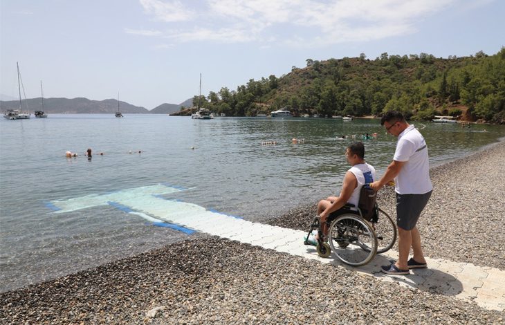 Bu fotoğraf, engelli bireylerin denize erişimini kolaylaştırmak amacıyla yapılmış özel bir yürüyüş yolunu gösteriyor. Sağ tarafta bir kişi, tekerlekli sandalyedeki bir bireyi kıyıya kadar getiriyor. Yol, tekerlekli sandalye ile rahat hareket edilebilecek şekilde denizin içine kadar uzanıyor. Arka planda tekneler, yüzen insanlar ve doğayla iç içe bir koy manzarası yer almakta. Fotoğraf, fiziksel engelleri aşmaya yönelik turizm altyapısının geliştiğini ve kapsayıcılığı temsil ediyor.