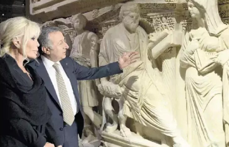 Bu fotoğraf, bir müze veya tarihi eser sergisinde çekilmiş görünüyor. İki kişi — bir kadın ve bir erkek — detaylı oymalarla süslenmiş büyük bir antik kabartmayı (muhtemelen bir lahit ya da rölyef) inceliyor. Erkek kişi eseri eliyle işaret ederek bir şeyler anlatıyor; kadın ise dikkatle dinliyor ve izliyor. Arka planda, klasik döneme ait insan figürleri ve süslemeler yer almakta. Görsel, tarihi ve kültürel mirasa duyulan ilgiyi yansıtıyor.