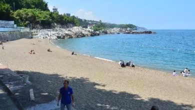 Bu fotoğraf, denize kıyısı olan geniş ve temiz bir plajı göstermektedir. Kumsal boyunca az sayıda insan güneşlenmekte veya denize girmektedir. Deniz oldukça durgun ve berraktır. Arka planda kayalık bir alan ve ağaçlarla kaplı bir tepe yer almakta. Sahilin kenarında bir yürüyüş yolu ve bazı yapılar da görülüyor. Ön planda ise birkaç kişi yürümekte veya plajı izlemektedir. Hava açık ve güneşlidir, yaz mevsimi izlenimi vermektedir.