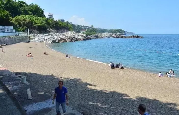 Bu fotoğraf, denize kıyısı olan geniş ve temiz bir plajı göstermektedir. Kumsal boyunca az sayıda insan güneşlenmekte veya denize girmektedir. Deniz oldukça durgun ve berraktır. Arka planda kayalık bir alan ve ağaçlarla kaplı bir tepe yer almakta. Sahilin kenarında bir yürüyüş yolu ve bazı yapılar da görülüyor. Ön planda ise birkaç kişi yürümekte veya plajı izlemektedir. Hava açık ve güneşlidir, yaz mevsimi izlenimi vermektedir.