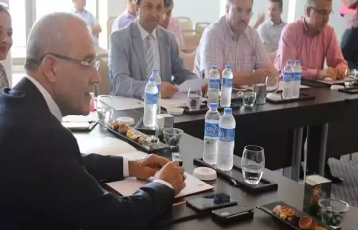Bu fotoğraf bir toplantı ortamını göstermektedir. Masalarda su şişeleri, not defterleri, cep telefonları, kalemler ve tabaklarda sunulmuş yiyecekler yer almaktadır. Ön planda takım elbiseli bir adam not alırken görülüyor. Arka planda ise başka kişiler de masada oturmakta ve toplantıya katılmaktadır. Ortam resmi ve profesyonel bir izlenim vermektedir.