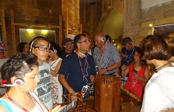 Fotoğrafta kapalı bir müze ya da sergi alanı görülüyor. Kalabalık bir grup ziyaretçi, ahşap maketlerin sergilendiği bir masanın etrafında toplanmış. Ziyaretçilerin çoğu kulaklık ve sesli rehber cihazları kullanıyor; bu da bir rehber eşliğinde gezi yaptıklarını gösteriyor. Masanın üzerinde tarihi sur, kule ve mancınık gibi yapıları temsil eden detaylı ahşap maketler bulunuyor. Arka planda taş duvarlar, kemerli geçitler ve sergi panoları görülüyor. Ortam sıcak tonlarda aydınlatılmış ve tarihi bir atmosfer hakim. Ziyaretçiler ilgiyle sergilenen objeleri inceliyor.