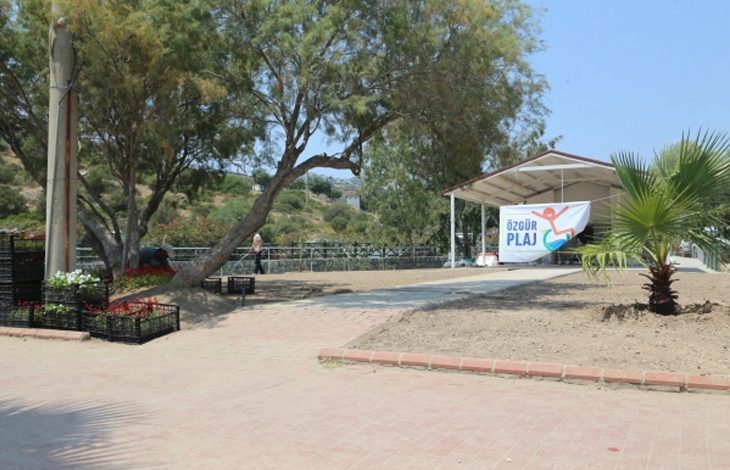 Bu fotoğraf, açık alanda çekilmiş ve “Özgür Plaj” tabelasının yer aldığı bir sahneyi göstermektedir. Sağ tarafta, üstü kapalı bir alanın girişinde büyük bir “Özgür Plaj” pankartı asılmış. Sol ve arka kısımlarda ağaçlar ve peyzaj düzenlemeleri görülüyor. Zeminde taş döşeme yollar, yanlarda çiçek kasaları ve yeni ekilmiş alanlar yer almakta. Görünen alan, engelli bireyler için erişilebilir şekilde düzenlenmiş bir plaj alanının girişini temsil ediyor.