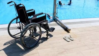 Fotoğrafta bir yüzme havuzu kenarı görülüyor. Ön planda boş bir tekerlekli sandalye ve hemen yanında beyaz bir çift terlik yer alıyor. Havuzun kenarında ayrıca engelli bireylerin havuza rahatça girebilmesi için tasarlanmış bir havuz asansörü (lift sistemi) bulunuyor. Havuzun içinde ise birkaç kişi suyun keyfini çıkarıyor. Hava güneşli ve açık; ortam temiz ve bakımlı görünüyor. Bu görüntü, engelli bireylerin erişimine uygun, engelsiz bir tatil tesisini yansıtıyor.