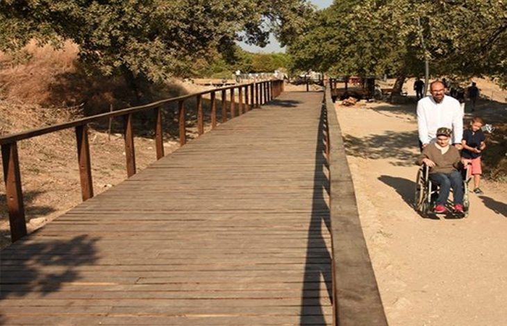 Fotoğrafta doğa içerisinde yer alan bir yürüyüş yolu görülüyor. Sol tarafta, ahşap malzemeden yapılmış, yanlarında korkulukları bulunan geniş ve düz bir yürüyüş platformu bulunuyor. Sağ tarafta ise toprak bir patika uzanıyor. Patika üzerinde bir adam, tekerlekli sandalyedeki bir bireyi itiyor; yanlarında küçük bir çocuk da yürüyor. Yolun iki yanında ağaçlar gölge oluşturmuş. Havanın güneşli ve sıcak olduğu anlaşılıyor. Ortam sakin, doğal ve erişilebilir bir gezi yolunu yansıtıyor.