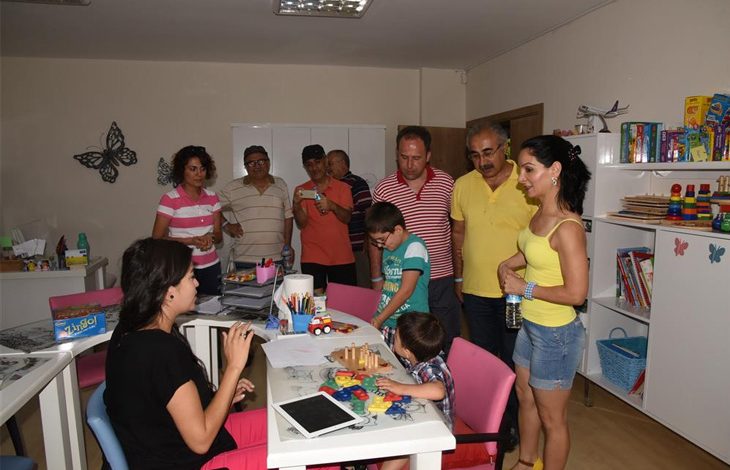 Bu fotoğraf bir sınıf ya da danışma odasını göstermektedir. Ön planda bir masa etrafında oturmuş ve oyuncaklarla oynayan iki çocuk yer almakta. Masanın bir ucunda siyah tişörtlü bir kadın oturuyor ve çocuklarla ilgileniyor. Arka planda ise birkaç yetişkin ayakta durmakta, bazıları çocukları izliyor, bazıları telefonla fotoğraf çekiyor. Oda rengârenk, raflarda kitaplar ve oyuncaklar bulunuyor.