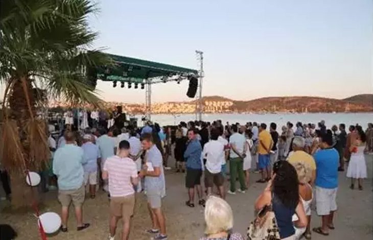 Fotoğrafta deniz kenarında düzenlenen bir etkinlik görülüyor. Ön planda ve orta alanda birçok insan sahilde toplanmış. Katılımcıların bazıları ayakta sohbet ediyor, bazıları ise sahnedeki etkinliği izliyor. Sol tarafta bir palmiye ağacı yer alıyor. Deniz kıyısında kurulan yeşil tente ile kaplı sahnede müzisyenler veya konuşmacılar bulunuyor. Sahnenin üstünde ışıklandırma ve ses sistemleri dikkat çekiyor. Arka planda deniz ve kıyıya paralel uzanan tepeler görülüyor. Hava açık ve gün batımı saatlerine yakın olduğu anlaşılıyor. Genel olarak yaz akşamında düzenlenen keyifli ve sosyal bir açık hava etkinliğini yansıtıyor.