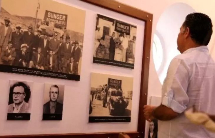 Fotoğrafta bir müze ortamı görülüyor. Sağ tarafta beyaz gömlek giymiş bir adam, duvara asılı bir pano üzerindeki siyah-beyaz fotoğraflara dikkatle bakıyor. Panoda, üzerinde "Sünger Kooperatifi" yazan eski dönem fotoğrafları bulunuyor. Fotoğraflar, geçmişteki süngercilik faaliyetlerini ve bu işle uğraşan insanları yansıtıyor. Ayrıca panoda iki adet portre fotoğrafı da yer almakta. Mekânın duvarı beyaz renkli ve duvarda yuvarlak bir pencere veya açıklık bulunuyor. Genel olarak tarihî ve kültürel bir atmosfer hakim.