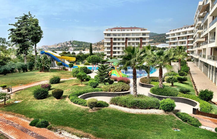 Fotoğrafta, Fortuna Resort’un havuz ve yeşil alanları var. Farklı boyutlarda ve uzunluklarda ağaçlar ile birlikte yeşil alanın dışında tatil yapılan binanın da dış bölümü görülmektedir.