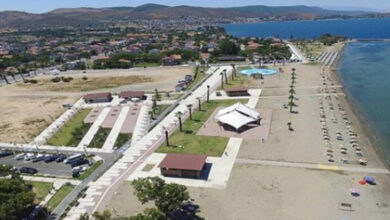 Yüksek bir yerden alınmış görüntü ile uzun bir sahil ve plajın görseli yer alıyor. Görselde uzak mesafede bazı evlerin olduğu da görülmektedir.