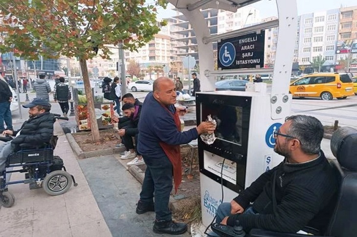 Engelli bireyler şarj istasyonunda araçlarını şarj ederken esnaf İdris Ekinci ise şarj istasyonunu silerek temizliyor.