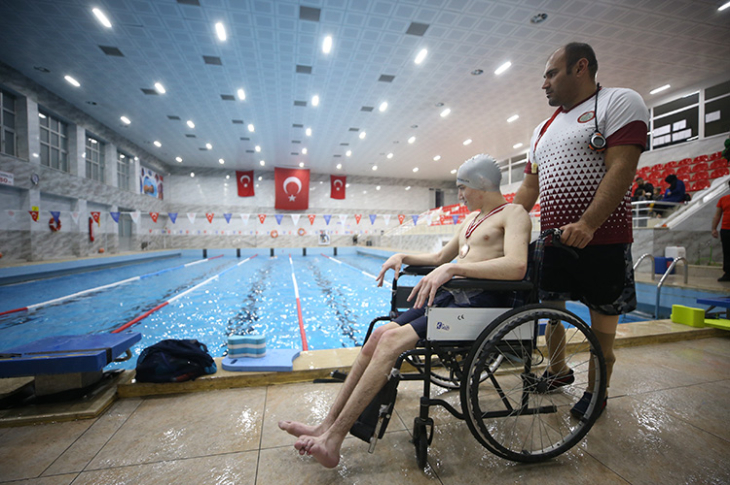 Öğretmen Ali Uzun, havuz salonunda öğrencisinin bindiği tekerlekli sandalyesini sürüyor.