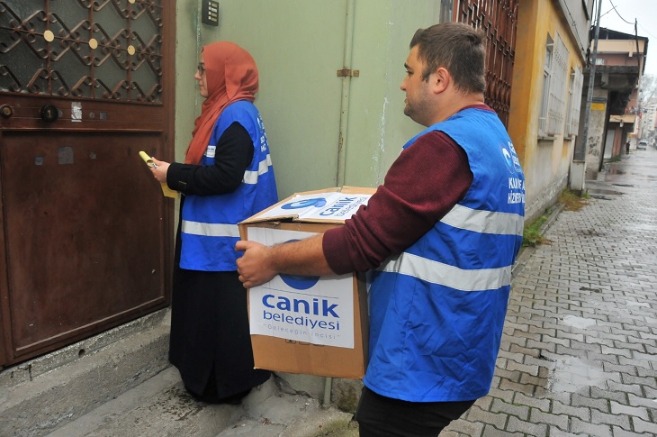 Bir ihtiyaç sahibi hanenin kapısına kumanya getiren 2 görevli objektife yansıyor.