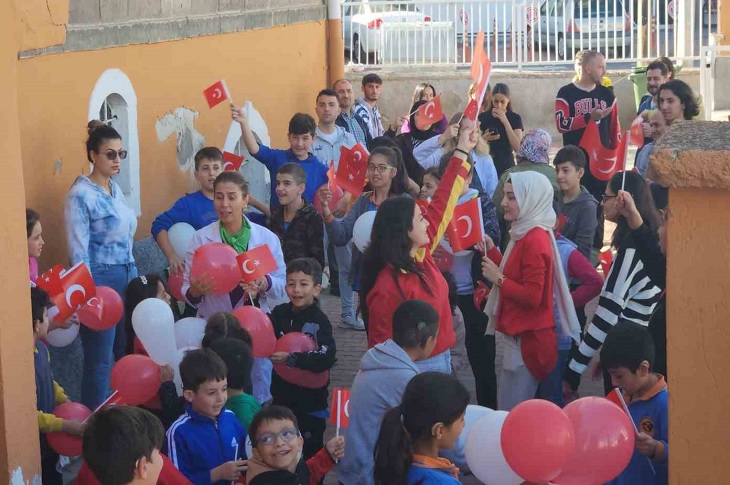 Okul bahçesinde ellerinde bayrak ve balon olan çok sayıda çocuk objektife yansıyor.