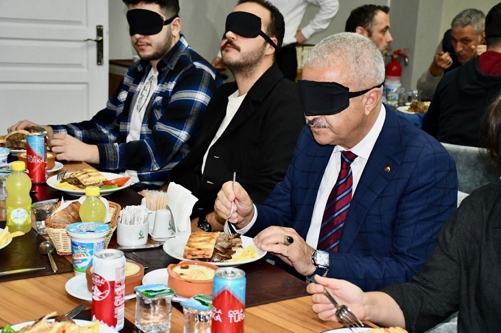 Yemek masalarında, gözleri kapalı halde yemek yemeye çalışan çok sayıda katılımcı objektife yansıyor.