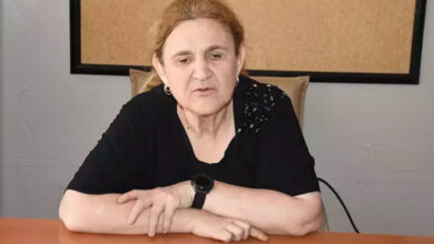 Figen Bilgen bir sandalyede oturup, kollarını masanın üstünde birleştirmektedir.