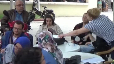 Kalabalık bir engelli grubunun masa etrafında toplanmış ve engelli bireylere maske dağıtılmaktadır.