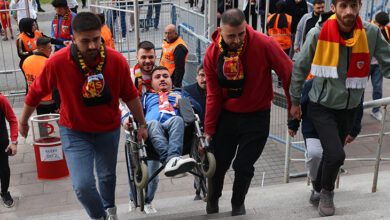 Hüseyin Deniz, tekerlekli sandalye üzerinde üç erkek taraftar tarafından stadyum merdivenlerinden yukarı doğru taşınıyor.