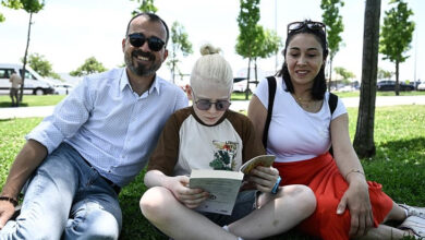 Parkta yeşil toprak üzerinde oturarak kitap okuyan albinizmli birey ve ailesi görselde var.