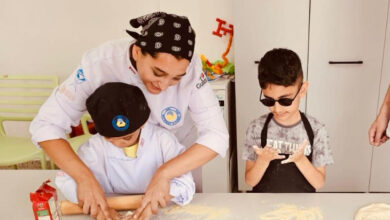 Görme engelli çocuklara, pizza hamuru açmaları için ellerinden ve oklavadan tutarak bir kadın öğrenci destek oluyor.