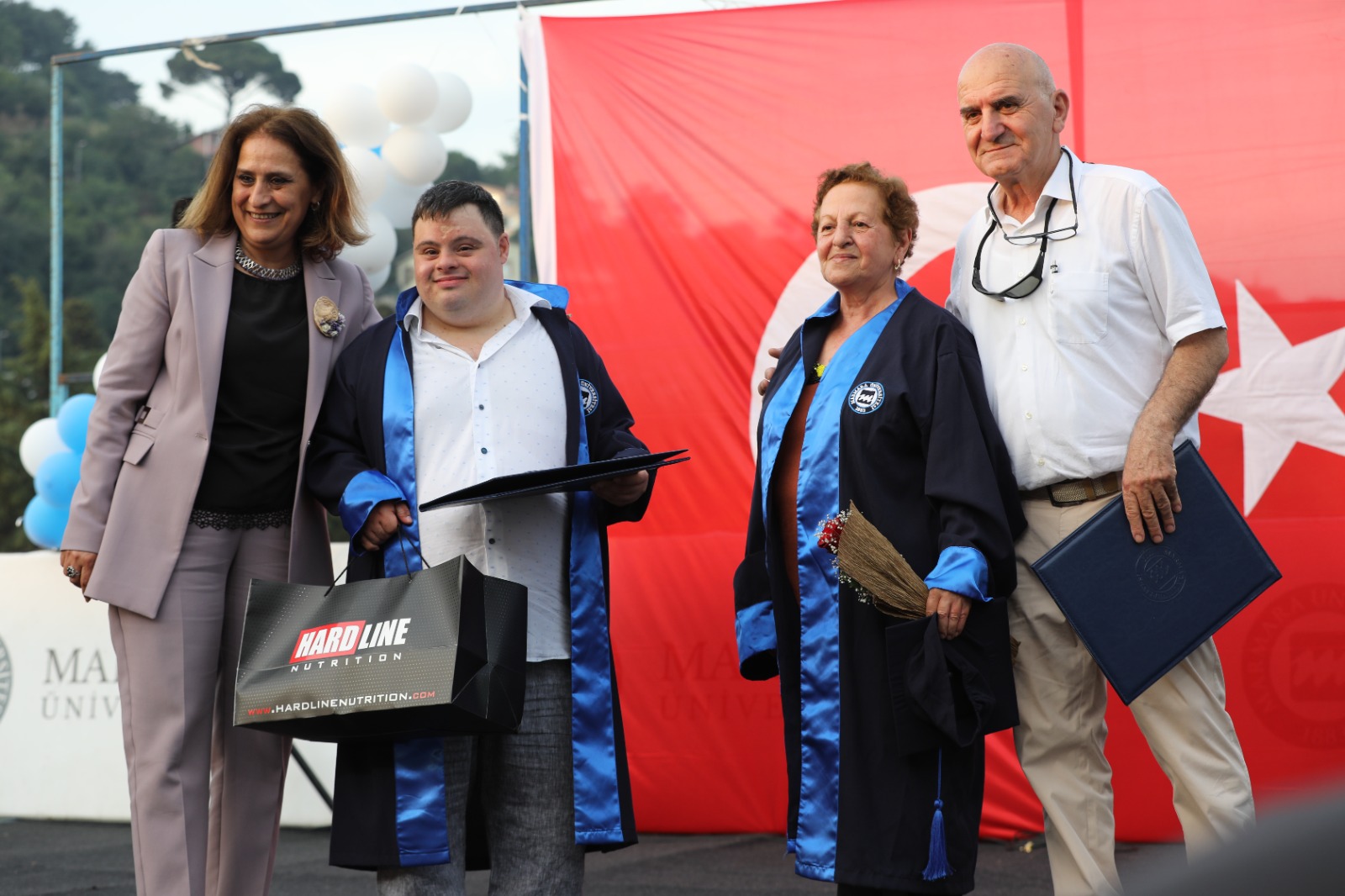 Down sendromlu Berk Canbulat'ın elinde diploması ve hediyesi, yanında ise üniversite hocası var. Anne Canbulat'a da cübbe giydirilmiş ve baba Canbulat'da yanında duruyor.