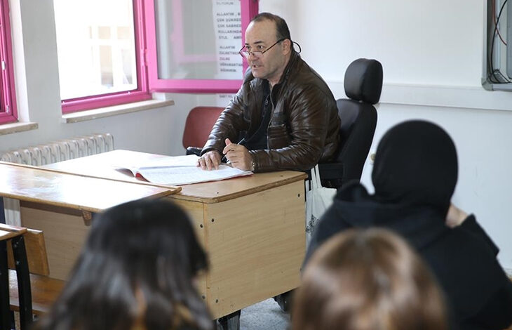 Akülü tekerlekli sandalye üzerinde oturan Bahattin Yıldırım, öğretmen masası arkasından öğrencilerine ders anlatıyor.