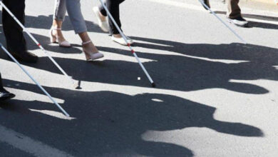Fotoğraf, bir grup insanın beyaz bastonlarla asfalt yolda yürürken çekilmiştir. Beyaz bastonla yürüyen kişilerin ayakları, beyaz bastonları ve yere yansıyan gölgeleri var.