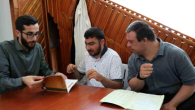 Masa etrafında oturmuş engelli bireyler ve cami imamı önlerindeki kitaplarla ilgili bir şeyler konuşuyorlar.