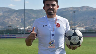 İşitme engelli futbolcu Emre Kılınç, futbol sahasında bir elinde futbol topu tutuyor ve diğer el baş parmağını yukarı doğru kaldırarak fotoğraf çekilmektedir.