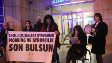 Fotoğrafta, tekerlekli sandalye kullanıcısı engelli bir kadın elindeki kağıttan bakarak ve diğer elindeki megafona konuşuyor. Diğer engelli bireylerde pakart tutuyor, pankartta "ENGELLİ ÇALIŞANLARA UYGULANAN MOBİNG VE AYRIMCILIK SON BULSUN" yazıyor.