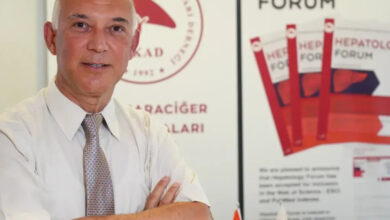 Türk Karaciğer Araştırmaları Derneği (TKAD) Başkanı Prof. Dr. Abdullah Zeki Karasu, dernek logosu önünde hafif yan durarak ve kollarını birbirine bağlayarak fotoğraf için poz veriyor.