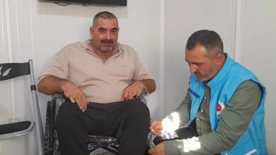 Engelli birey hediye edilen tekerlekli sandalye üzerinde oturuyor ve müftülük görevlisi sandalyenin poşetlerini çıkarıyor.