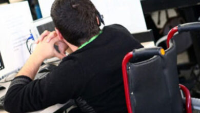 Tekerlekli sandalye üzerinde bulunan engelli birey masa başında bilgisayar ile çalışıyor.