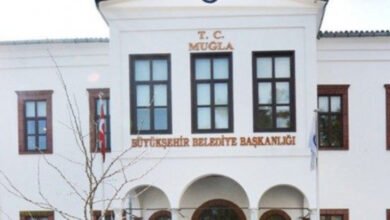 Beyaz renkli bir bina üzerinde "T.C MUĞLA BÜYÜKŞEHİR BELEDİYE BAŞKANLIĞI" yazısı var.
