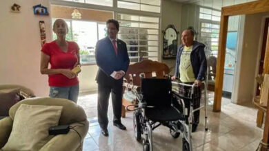 Venezuelalı engelli birey, iki ayaklı demir yapımı yürüteci tutarak ayakta duruyor. İstanbul Milletvekili Serkan Bayram akülü tekerlekli sandalyenin arkasında hafif dönerek yanındaki büyükelçinin söylediklerini dinliyor. Fotoğrafın en sol tarafında ise Caracas Büyükelçisi Aydan Karamanoğlu var.