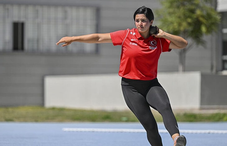 Paralimpik sporcu, gülle atışı için pozisyon alıyor. Paralimpik kadın sporcu, gülleyi boynunun altında eliyle tutuyor ve diğer elini omuz hizasının biraz üzerinde tutarak geriye doğru vücudunu çekiyor.