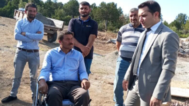 Tekerlekli sandalye kullanıcısı engelli muhtar Halil Yerlikaya, Kaymakam Mustafa Kartal ile karşılıklı olarak konuşuyor. Muhtar ve Kaymakamın çevresinde ise köy halkı konuşmaları dinliyor.