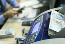 Para sayma makinesinin üzerinde 100 TL'lik banknotlar var. Fotoğrafta, flu bir şekilde hesap makinesi, 100 TL'lik banknotlar, bilgisayar ekranı ve klavyesi, masanın üzerinde duruyor. Aynı zamanda yüzü gözükmeyen bir kişi elindeki paraları sayıyor.